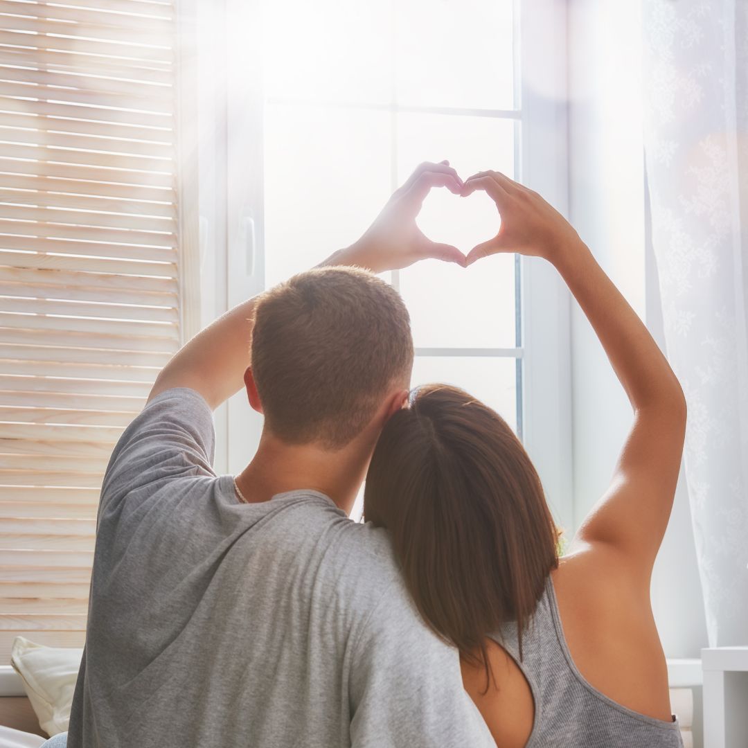زوجان من الخلف يرفعان أيديهما ويشكّلان قلبًا أمام نافذة مضيئة