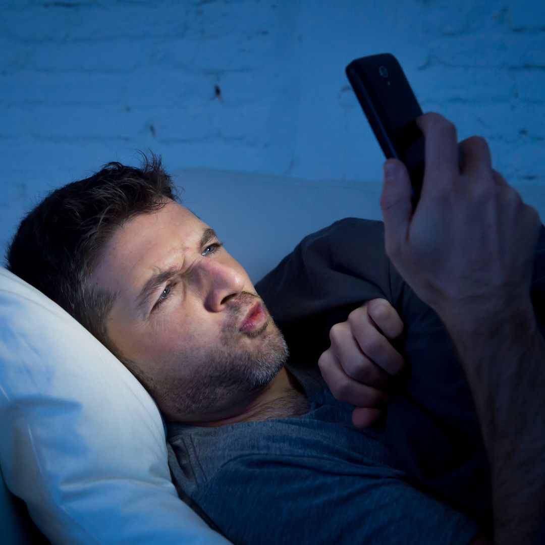 رجل مستلقٍ في السرير ليلاً ووجهه مضاء بشاشة هاتفه المحمول